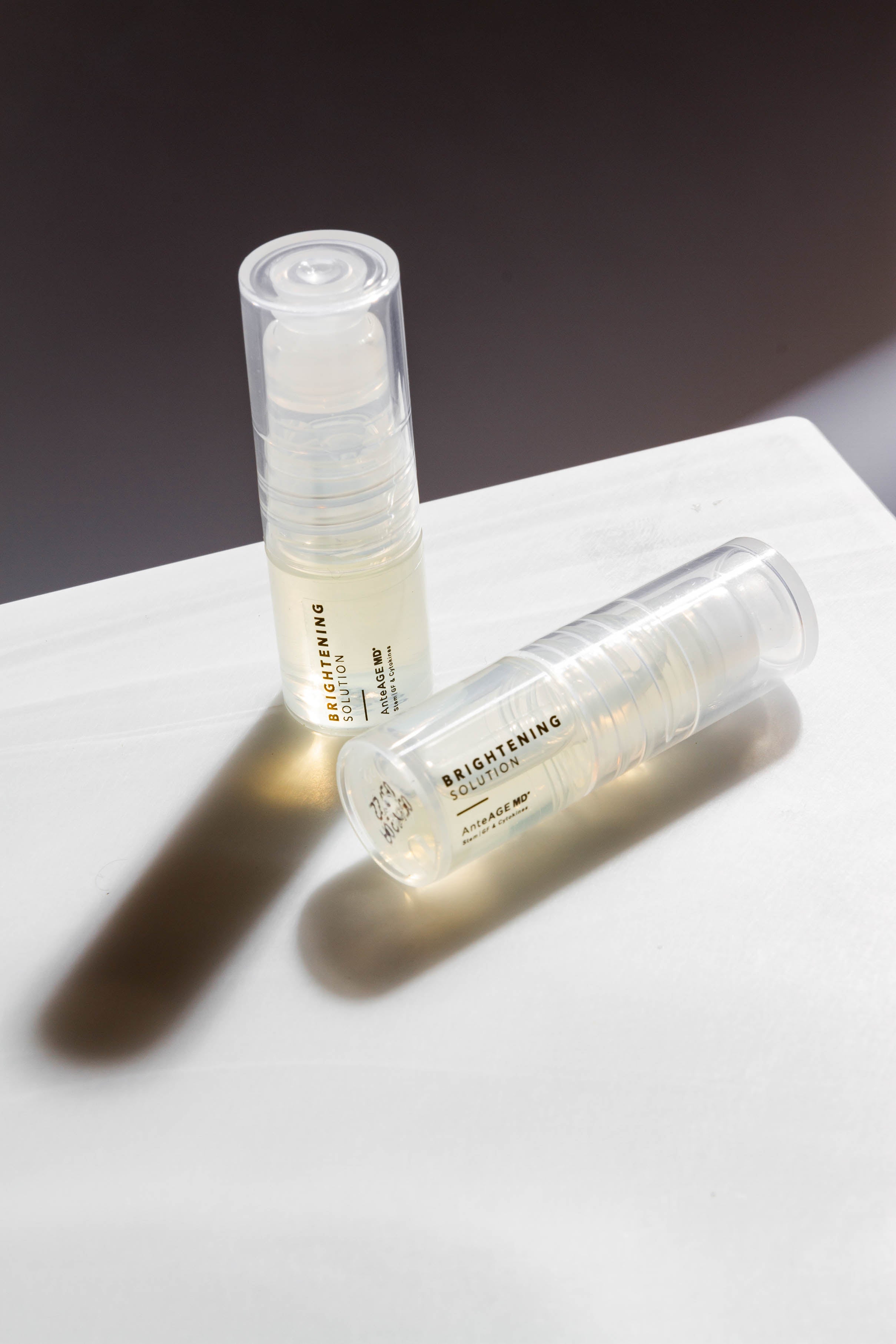 Two skincare serum bottles labeled 'Brightening' on a marble surface with a dark background.