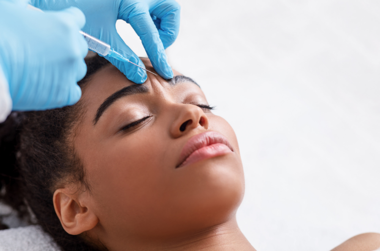 A close-up of a woman receiving a brow or eyebrow injection, with a practitioner's gloved hand holding a syringe near her eyebrow. The woman has closed eyes and relaxed skin, and the background is a clean, bright white.