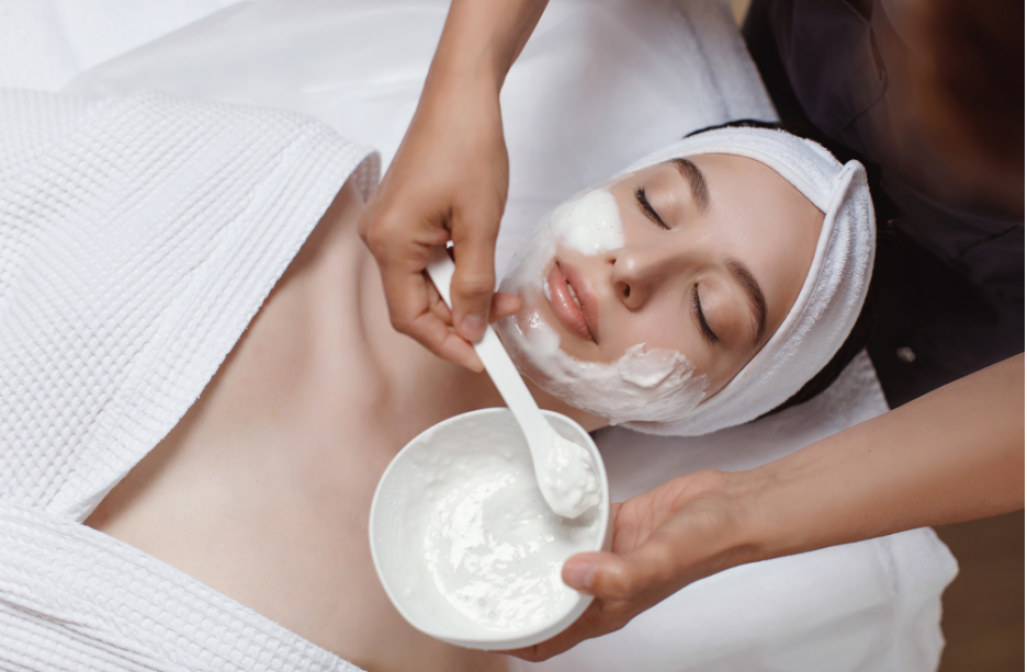 A woman lying down with a white head wrap and face mask while a spa worker applies a white facial cream from a small bowl with a spoon.