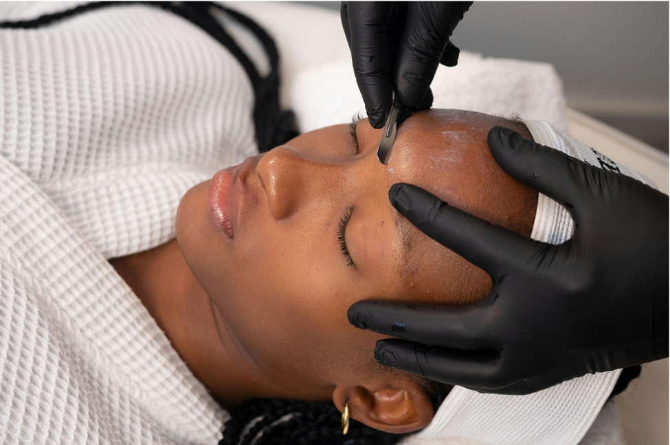 A close-up of a woman lying down with her eyes closed while a person wearing black gloves applies a small cosmetic tool to her forehead. She is wrapped in a white textured blanket, and the background is softly blurred.