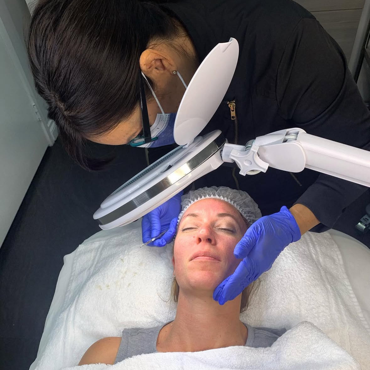 Person receiving a facial treatment with a professional using a magnifying lamp.