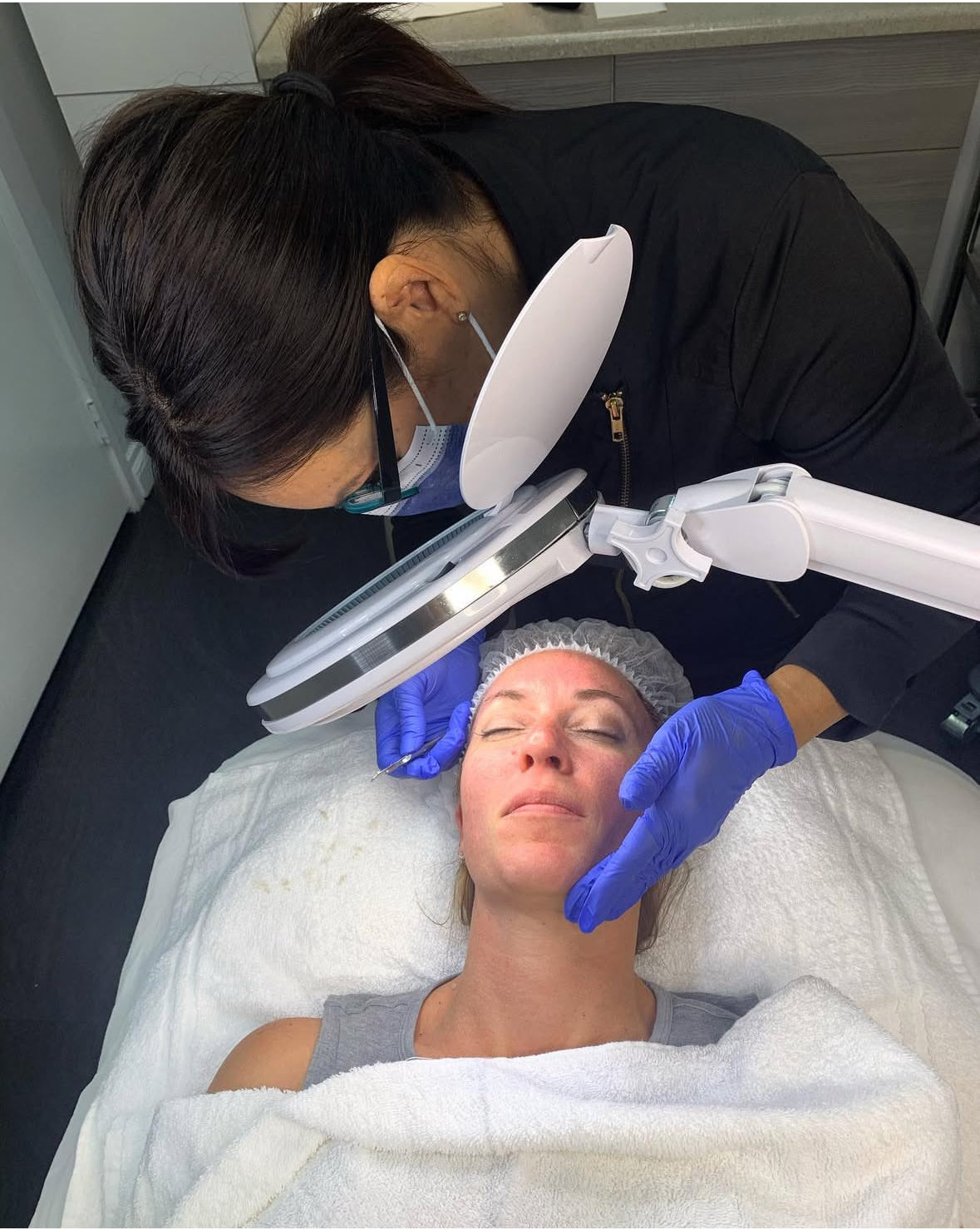 Person receiving a facial treatment with a professional using a magnifying lamp.