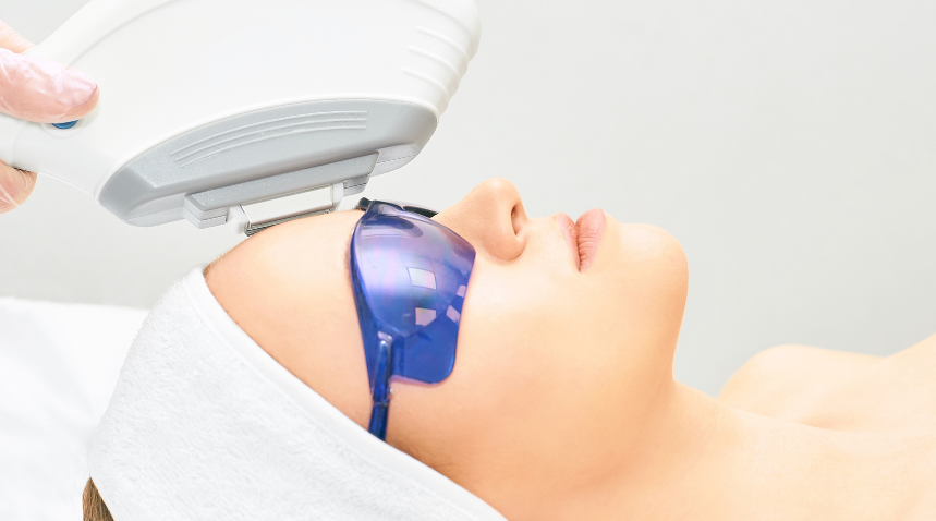 A close-up of a person lying down with a white headband, wearing blue protective glasses while a white handheld beauty device is being used on their forehead by a gloved hand.