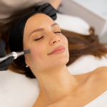 A woman with closed eyes lies on a treatment bed while a spa professional in black gloves applies a cosmetic product to her cheek with a small handheld applicator. She wears a black headband and a white towel is draped over her shoulder in a bright, clean beauty setting.